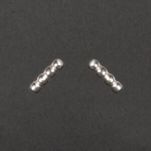 Könnte beinhalten: Ein Paar silberne Ohrstecker. Jeder Ohrstecker besteht aus fünf kleinen, verbundenen, abgerundeten Kugeln. Die Ohrringe sind vor einem dunkelgrauen Hintergrund platziert, der ihren metallischen Glanz und ihr schlichtes Design hervorhebt.