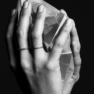 May include: Black and white image of hands holding a large, faceted, clear crystal. Two thin, silver rings adorn the fingers. The crystal reflects light, creating a play of shadows and highlights. The background is a solid black.