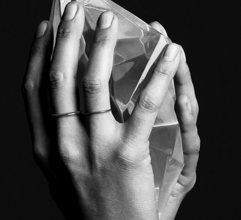 May include: Black and white image of hands holding a large, faceted, clear crystal. Two thin, silver rings adorn the fingers. The crystal reflects light, creating a play of shadows and highlights. The background is a solid black.