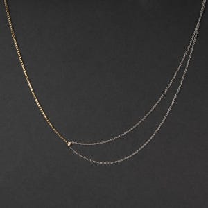May include: A two-strand necklace featuring a gold chain on the left and a silver chain on the right. The gold chain is a box chain, and the silver chain is a delicate link chain. A small rectangular charm joins the two chains.