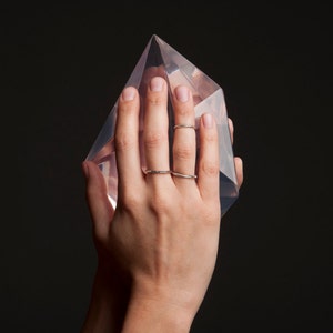 May include: A clear, geometric crystal sculpture is held in a hand, with two silver rings on the fingers. The crystal is a transparent, multi-faceted shape, reflecting light. The background is a solid black.