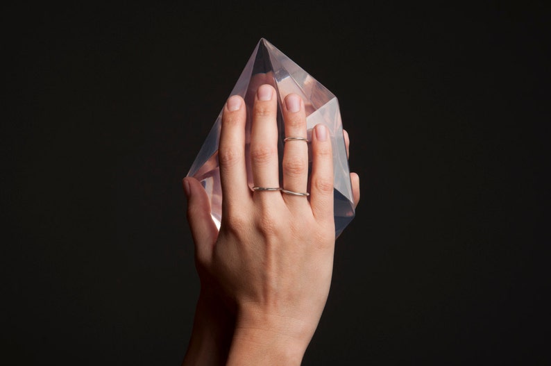 May include: A clear, geometric crystal sculpture is held in a hand, with two silver rings on the fingers. The crystal is a transparent, multi-faceted shape, reflecting light. The background is a solid black.