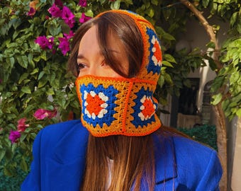 May include: A colorful crochet balaclava in orange, blue, and white. The balaclava is made of square crochet patches and is worn with a blue blazer. The background features green foliage and pink flowers.