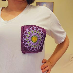 Puede incluir: Camiseta blanca con cuello en V con un bolsillo de ganchillo. El bolsillo es cuadrado y presenta un diseño floral con colores morado, verde y amarillo. El trabajo de ganchillo es detallado y añade un toque artesanal a la camiseta.