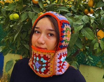 May include: A colorful, crocheted balaclava in a granny square pattern. The balaclava features a mix of red, orange, blue, yellow, and purple yarn. The head covering is designed to cover the head and neck, with an opening for the face.
