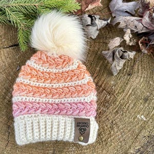 May include: A handmade crochet beanie in a gradient of colors, including cream, pink, and orange, topped with a fluffy cream pom-pom. The hat features a textured stitch pattern and a leather tag that reads "Campfire Crochet".