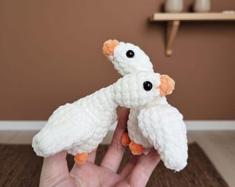 May include: Two white crochet geese with orange beaks and feet, and black eyes. The geese are handmade and appear soft and plush. The background is a solid brown color.