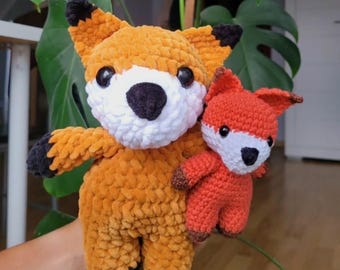 May include: Two crocheted fox stuffed animals. The larger fox is orange with a white muzzle, black ears, and black paws. The smaller fox is also orange with a white muzzle and brown paws. The foxes are handmade.