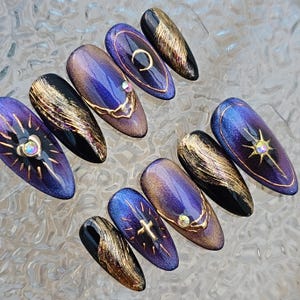 May include: A collection of ten long, oval-shaped artificial nails. Each nail displays a dark purple and gold colour scheme with diverse designs, including gold accents, starbursts, and a cross. Some nails have a black base.