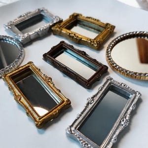 May include: A collection of seven small mirrors in various shapes and finishes. The set includes rectangular mirrors with ornate frames in silver, gold, and dark brown, alongside two round mirrors with beaded edges in silver and gold tones.