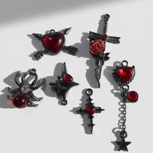 May include: A collection of six dark metal charms featuring red accents. The set includes a heart pierced by arrows, a sword with a red rose, a spider with a red bead, a four-pointed star with a red gem, a cross with a red heart, and a heart pendant with a chain and star.