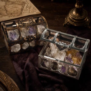 Puede incluir: Dos cofres del tesoro arqueados con marcos metálicos y paneles transparentes, uno con acabado en bronce antiguo y otro en plata pulida. Ambos cofres están llenos de cristales minerales en bruto, incluyendo cuarzo transparente, amatista y citrino.