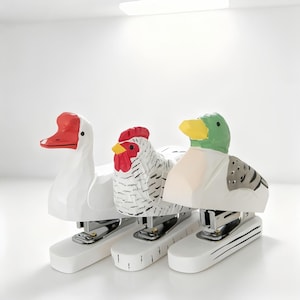 May include: Three whimsical staplers shaped like a duck, a chicken, and a goose. Each stapler has a white base with black stripes and a colorful animal head. The duck has a green head and yellow beak.