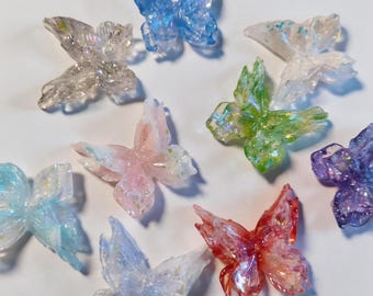 May include: A collection of nine butterfly-shaped ornaments crafted from translucent, iridescent resin. Each butterfly features a unique colour, including shades of blue, pink, green, red, purple, and clear, all embedded with fine, shimmering glitter particles.