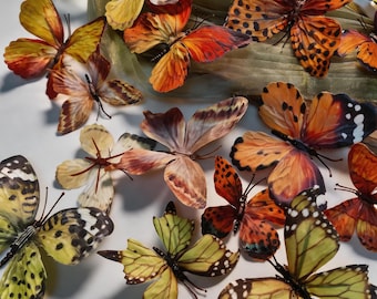 May include: A collection of decorative artificial butterflies in various sizes and colors, including shades of yellow, orange, brown, and black. Each butterfly features detailed wing patterns, thin wire antennae, and delicate, textured fabric wings.