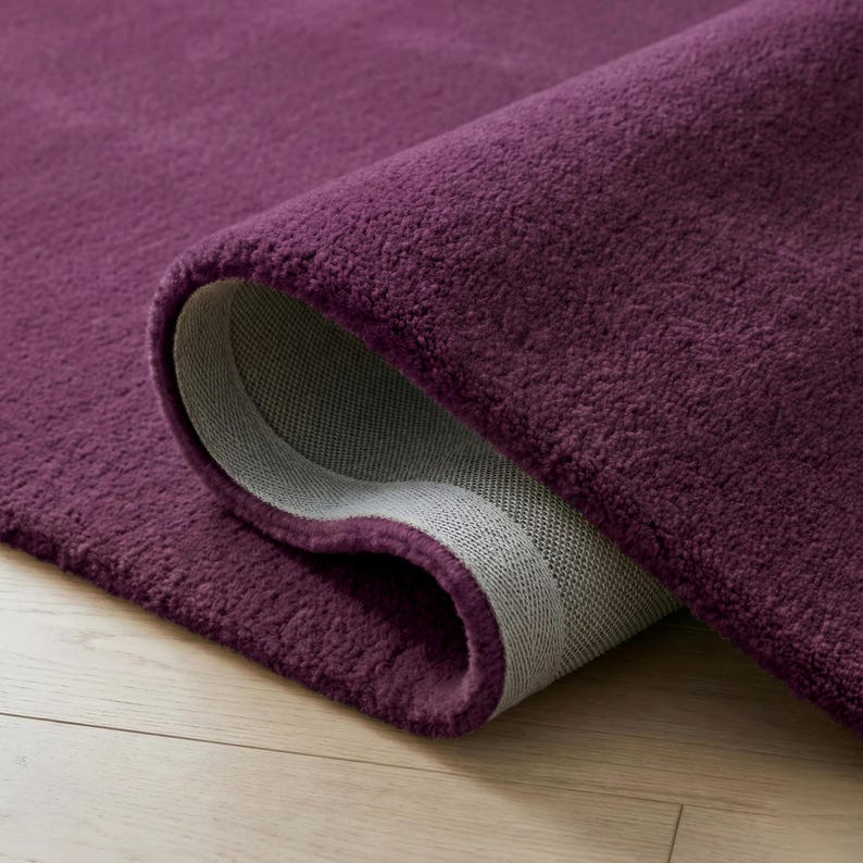 May include: A rolled-up, plush, purple rug with a soft, textured surface. The underside of the rug is a light gray color, revealing a woven backing. The rug is displayed on a light-colored wooden floor.