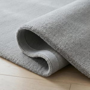 May include: A rolled-up, light gray rug with a plush, textured surface. The rug's edge reveals a woven backing. The rug is on a light-colored wooden floor.