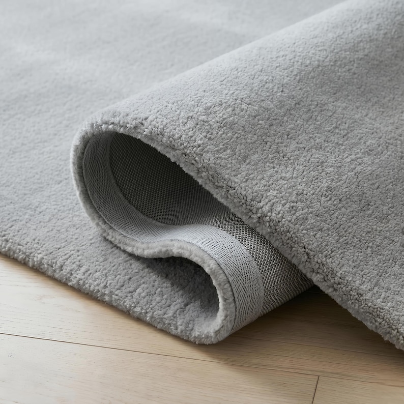 May include: A rolled-up, light gray rug with a plush, textured surface. The rug's edge reveals a woven backing. The rug is on a light-colored wooden floor.