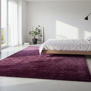 Pode incluir: Um tapete grande, retangular, roxo escuro domina o primeiro plano de um quarto luminoso. O tapete está posicionado sob uma cama com uma estrutura de madeira clara e um edredom branco. Uma planta em vaso e uma obra de arte emoldurada também são visíveis.
