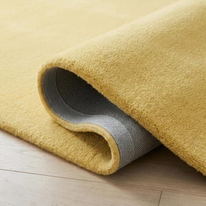 May include: A rolled-up, golden-yellow rug with a plush texture. The underside of the rug is a gray, woven material. The rug is on a light-colored wooden floor.