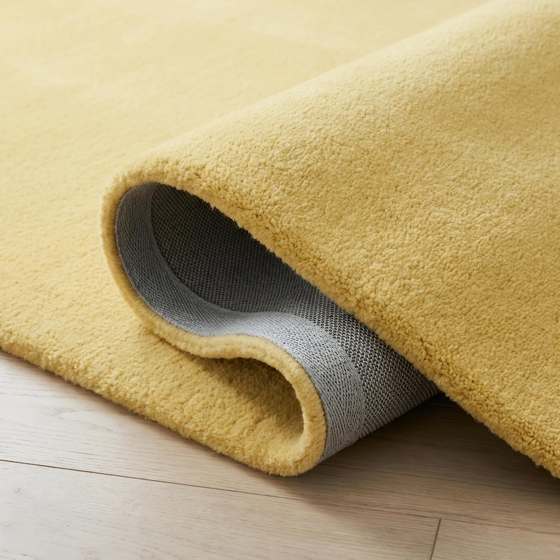 May include: A rolled-up, golden-yellow rug with a plush texture. The underside of the rug is a gray, woven material. The rug is on a light-colored wooden floor.