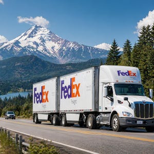 May include: A white FedEx Freight semi-truck with two trailers drives on a highway. The FedEx logo is visible in purple and orange. A snow-capped mountain and a lake are in the background.