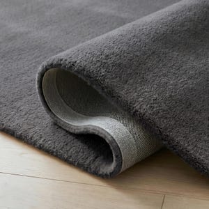 May include: A rolled-up, plush gray rug with a soft, textured surface. The underside of the rug is a lighter gray with a woven pattern. The rug is on a light-colored wooden floor.