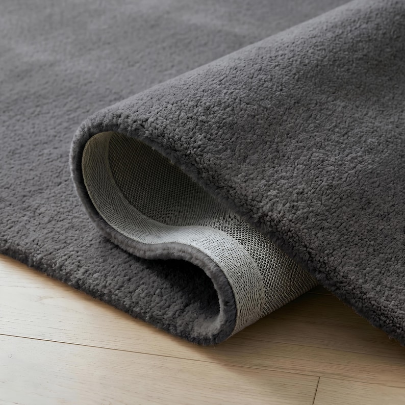 May include: A rolled-up, plush gray rug with a soft, textured surface. The underside of the rug is a lighter gray with a woven pattern. The rug is on a light-colored wooden floor.