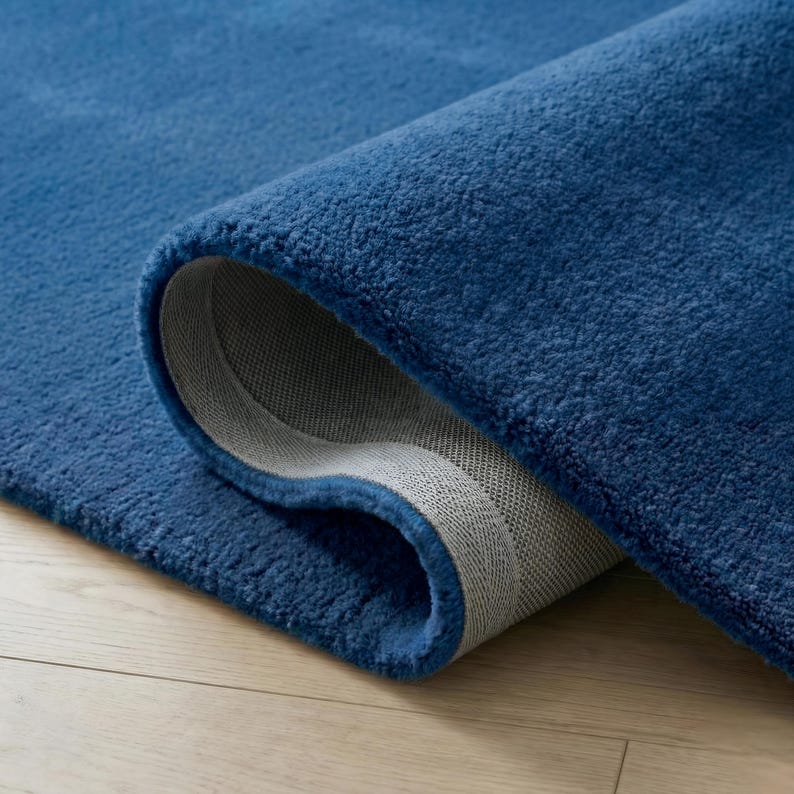 May include: A rolled-up, plush, blue rug with a soft, textured surface. The underside of the rug is a light gray color, and the edges are neatly finished. The rug is placed on a light-colored wooden floor.