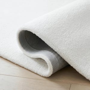 May include: A rolled-up white rug with a plush, textured surface. The underside of the rug is a gray woven material. The rug is on a light-colored wooden floor.