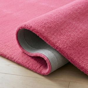 May include: A rolled-up, plush, pink rug with a gray backing. The rug's texture is soft and dense, with a close-up view highlighting the fibers and the rolled edge. The rug is on a light-colored wooden floor.