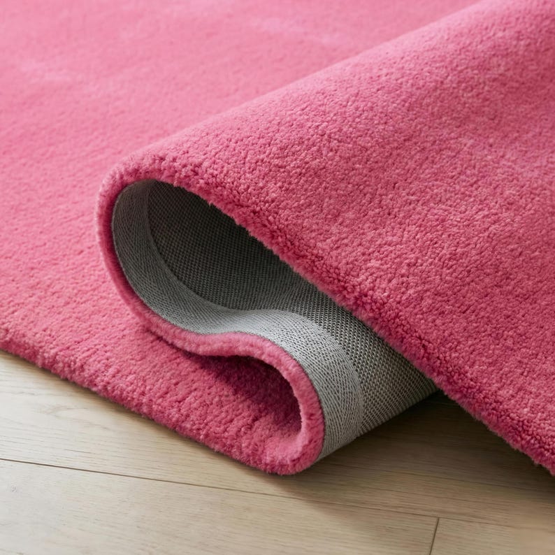 May include: A rolled-up, plush, pink rug with a gray backing. The rug's texture is soft and dense, with a close-up view highlighting the fibers and the rolled edge. The rug is on a light-colored wooden floor.