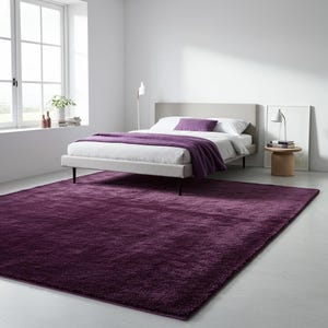 May include: A bedroom with a modern bed, grey headboard, white bedding, and a purple throw and pillow. A large, deep purple rug covers the floor. A window and side table are also visible. The rug is a deep shade of purple.