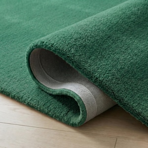 May include: A rolled-up, emerald green rug with a plush, textured surface. The underside reveals a gray, woven backing. The rug's edges are neatly finished, showcasing the quality of the material.
