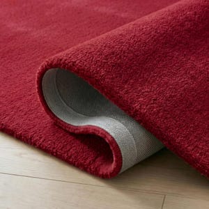 May include: A rolled-up, plush red rug with a visible gray backing. The rug's texture is dense and soft, with a deep red hue. The rug is partially rolled, revealing the underside.