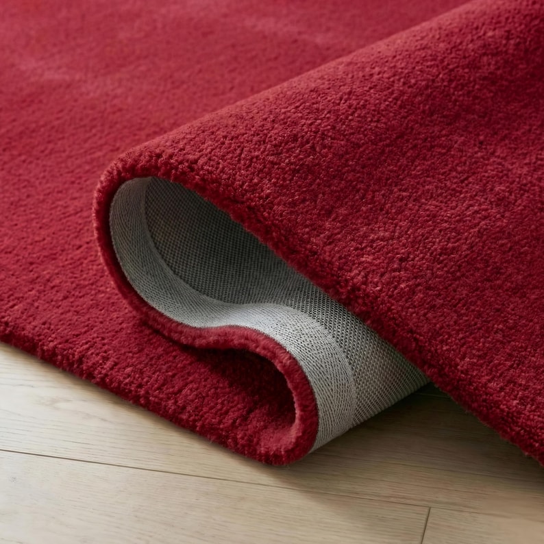 May include: A rolled-up, plush red rug with a visible gray backing. The rug's texture is dense and soft, with a deep red hue. The rug is partially rolled, revealing the underside.