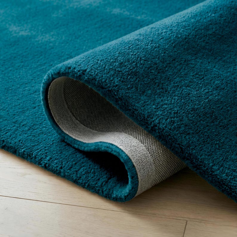 May include: A rolled-up, teal-colored rug with a plush, textured surface. The underside of the rug is visible, revealing a gray backing. The rug is placed on a light-colored wooden floor.