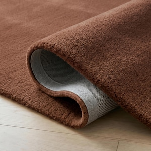 May include: A rolled-up, plush brown rug with a soft, textured surface. The underside of the rug is visible, showing a gray, woven backing. The rug is on a light-colored wooden floor.