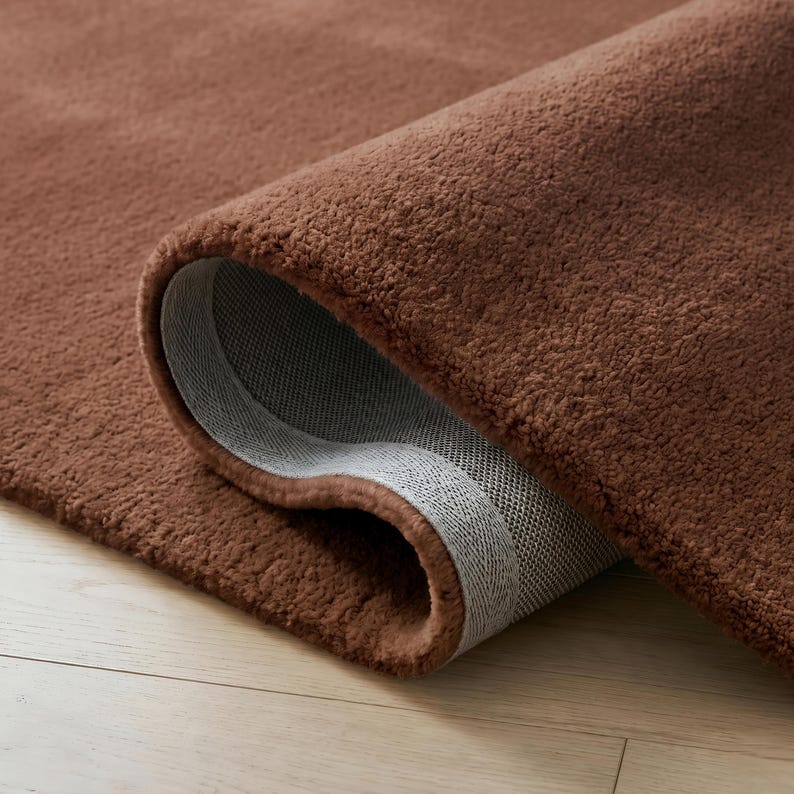 May include: A rolled-up, plush brown rug with a soft, textured surface. The underside of the rug is visible, showing a gray, woven backing. The rug is on a light-colored wooden floor.