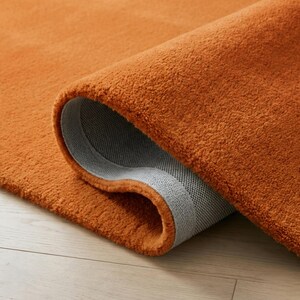 May include: Close-up of a rolled-up orange rug. The rug has a plush, textured surface and a gray backing. The color is a vibrant, warm orange, and the rug appears to be made of a soft material.