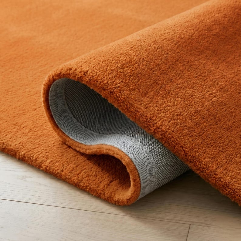 May include: Close-up of a rolled-up orange rug. The rug has a plush, textured surface and a gray backing. The color is a vibrant, warm orange, and the rug appears to be made of a soft material.