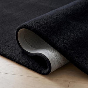 May include: A rolled-up, plush black rug with a soft, textured surface. The underside of the rug is a light gray color, revealing the woven backing. The rug is displayed on a light-colored wooden floor.