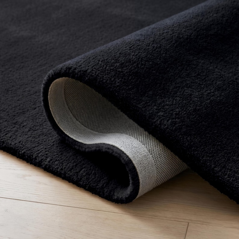 May include: A rolled-up, plush black rug with a soft, textured surface. The underside of the rug is a light gray color, revealing the woven backing. The rug is displayed on a light-colored wooden floor.