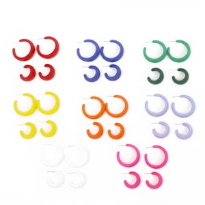 Op de afbeelding: Een collectie oorringen in diverse kleuren, waaronder rood, blauw, groen, donkergroen, geel, oranje, lichtpaars, wit en roze. Elke kleurset bevat twee verschillende maten ronde oorringen met metalen oorstekers.