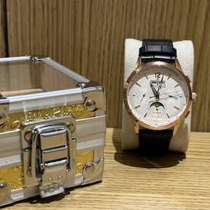 May include: A rose gold wristwatch with a white face and black leather band. The watch has multiple dials and a moon phase indicator. A silver and gold decorative box is to the left of the watch.