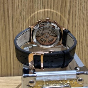 May include: Rose gold-toned wristwatch with a black leather strap. The watch features a transparent back, showcasing the intricate internal mechanisms. The clasp is also rose gold, and the watch is displayed on a metallic surface.