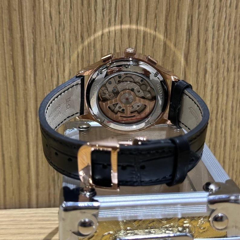 May include: Rose gold-toned wristwatch with a black leather strap. The watch features a transparent back, showcasing the intricate internal mechanisms. The clasp is also rose gold, and the watch is displayed on a metallic surface.