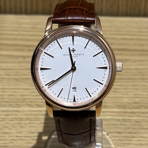 May include: A luxury wristwatch with a white face, gold-tone case, and brown leather band. The watch features a date display at the 6 o'clock position and the brand name "VACHERON CONSTANTIN NEVE" on the face. The watch is displayed on a clear stand.