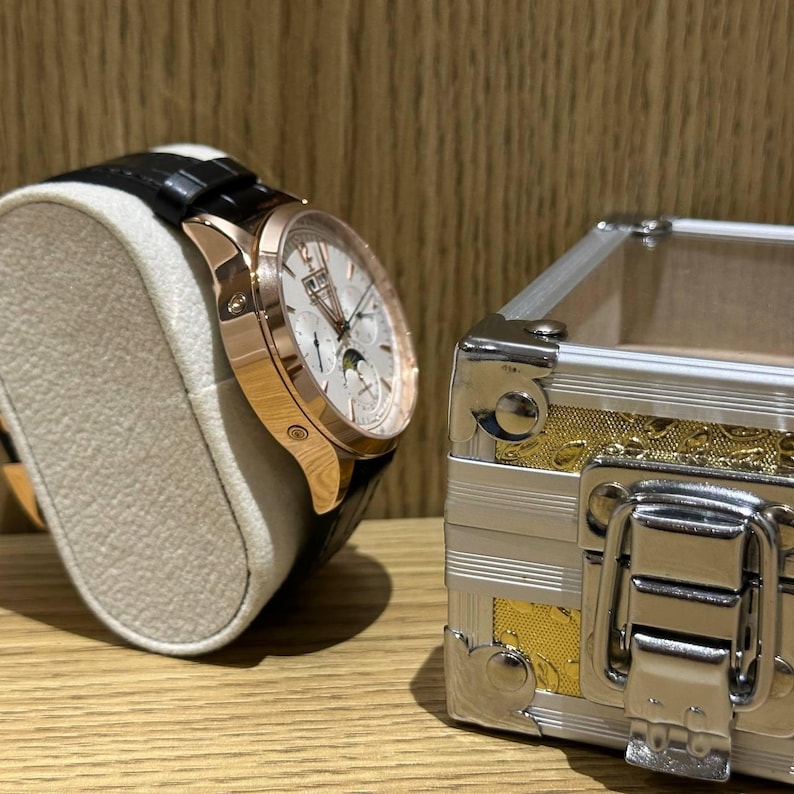 May include: A rose gold wristwatch with a black leather band rests on a beige display stand. The watch face is white with multiple dials. A silver and gold decorative box is to the right.