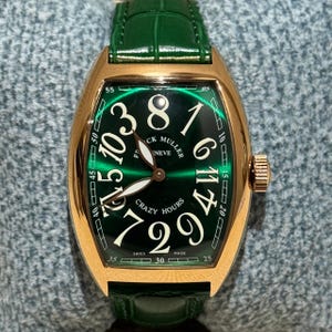 May include: A gold-toned wristwatch with a green face and a green leather strap. The watch face features large white numerals and the words "CRAZY HOURS" and "SWISS MADE". The watch has a unique curved rectangular shape.